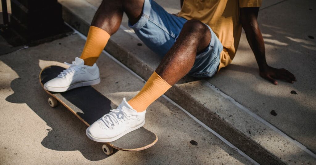 Crop anonymous African American male skater in t shirt and denim shorts sitting on sidewalk with feet on skateboard